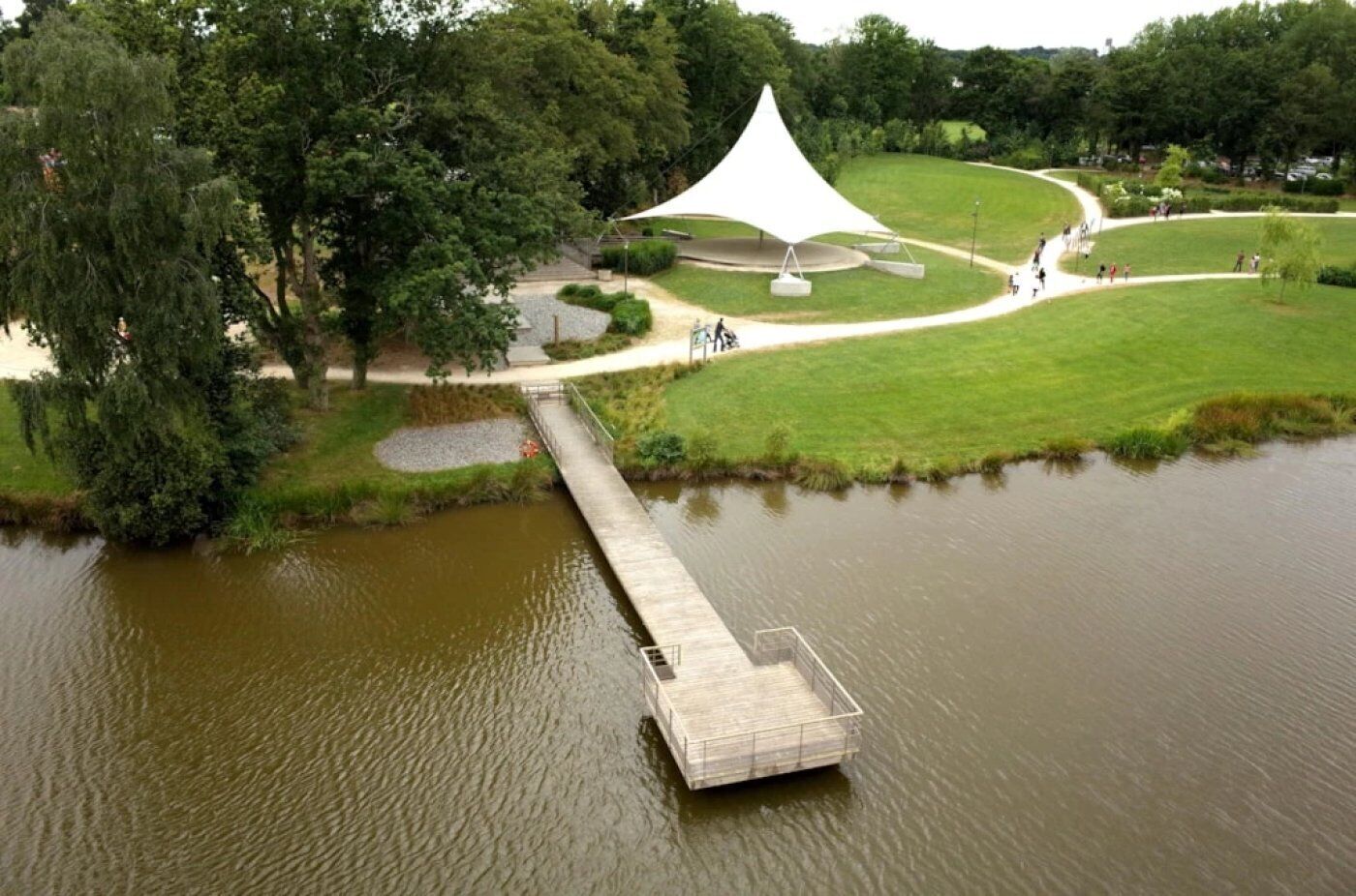 A park with a wooden bridge over a pond, a white tent-like structure, and a curved walking path.