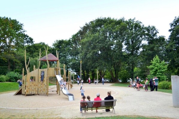 Un parque infantil con tobogán y estructuras para escalar, rodeado de árboles y una zona de arena.