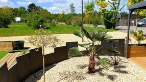 A landscaped garden with a palm tree, gravel area, wooden deck, and a modern building in the background. - ZEcamping 3* toboggans animaux acceptés cottage TV BBQ 30m2 4 pers (Loudéac)
