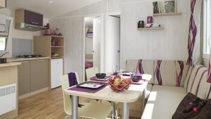 A compact kitchen with a dining area, a sofa, and a small table with a basket of fruit. - ZEcamping 3* toboggans animaux acceptés cottage TV BBQ 30m2 4 pers (Loudéac)
