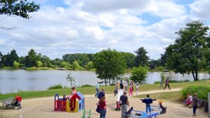 A playground with various play structures, a sandy area, and a lake in the background.