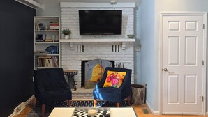 A living room with a white brick fireplace, a flat-screen TV mounted above it, a bookshelf, two armchairs, and a door. - Beautiful Bray Cottage on the Vineyard (West Cape May)