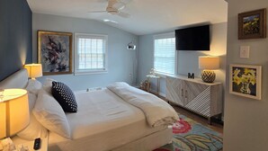 A bedroom with a bed, bedside lamps, a ceiling fan, a TV, and artwork on the walls. - Beautiful Bray Cottage on the Vineyard (West Cape May)