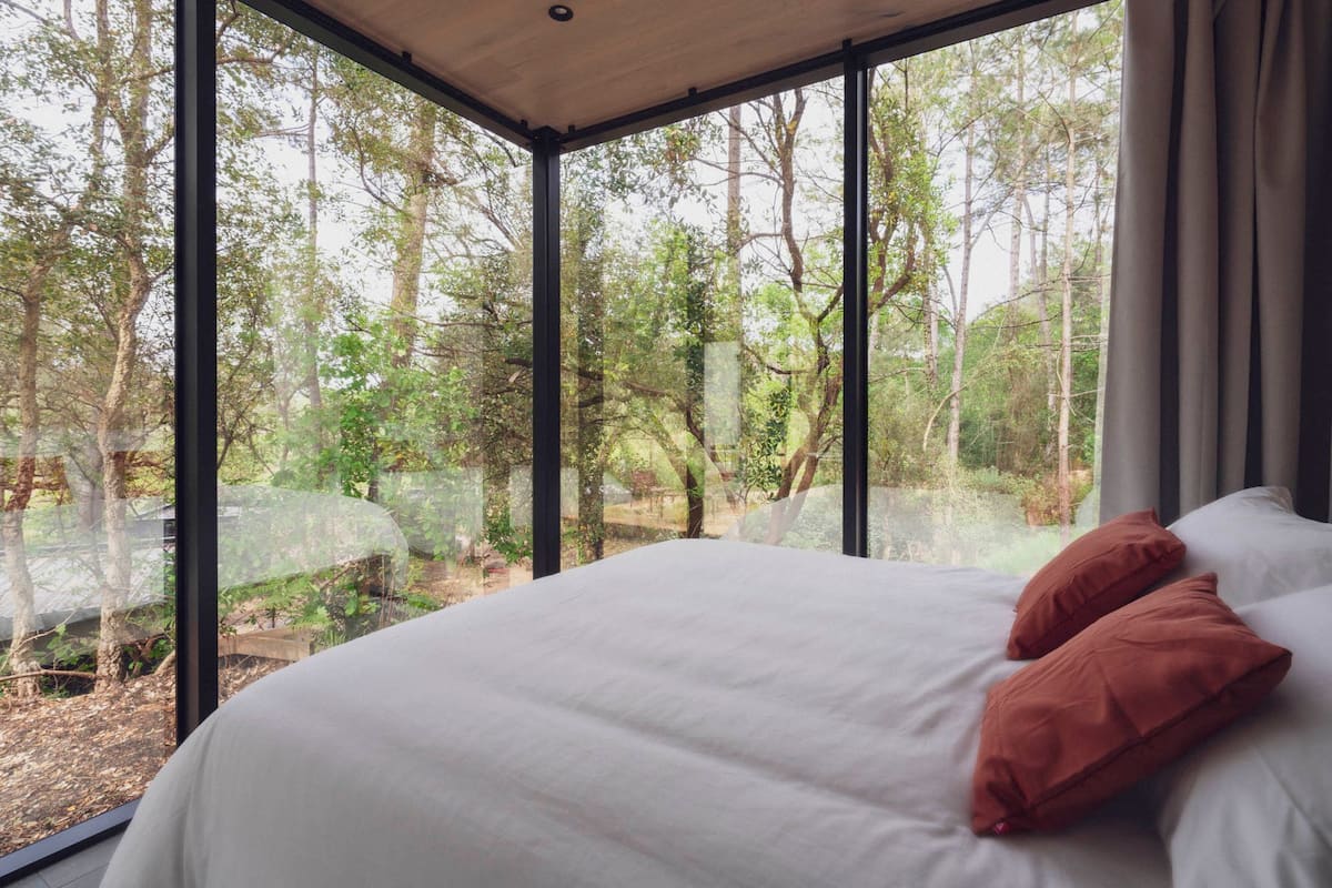 A bedroom with a large window, a bed with white linens and two pillows, and a view of a forested area.