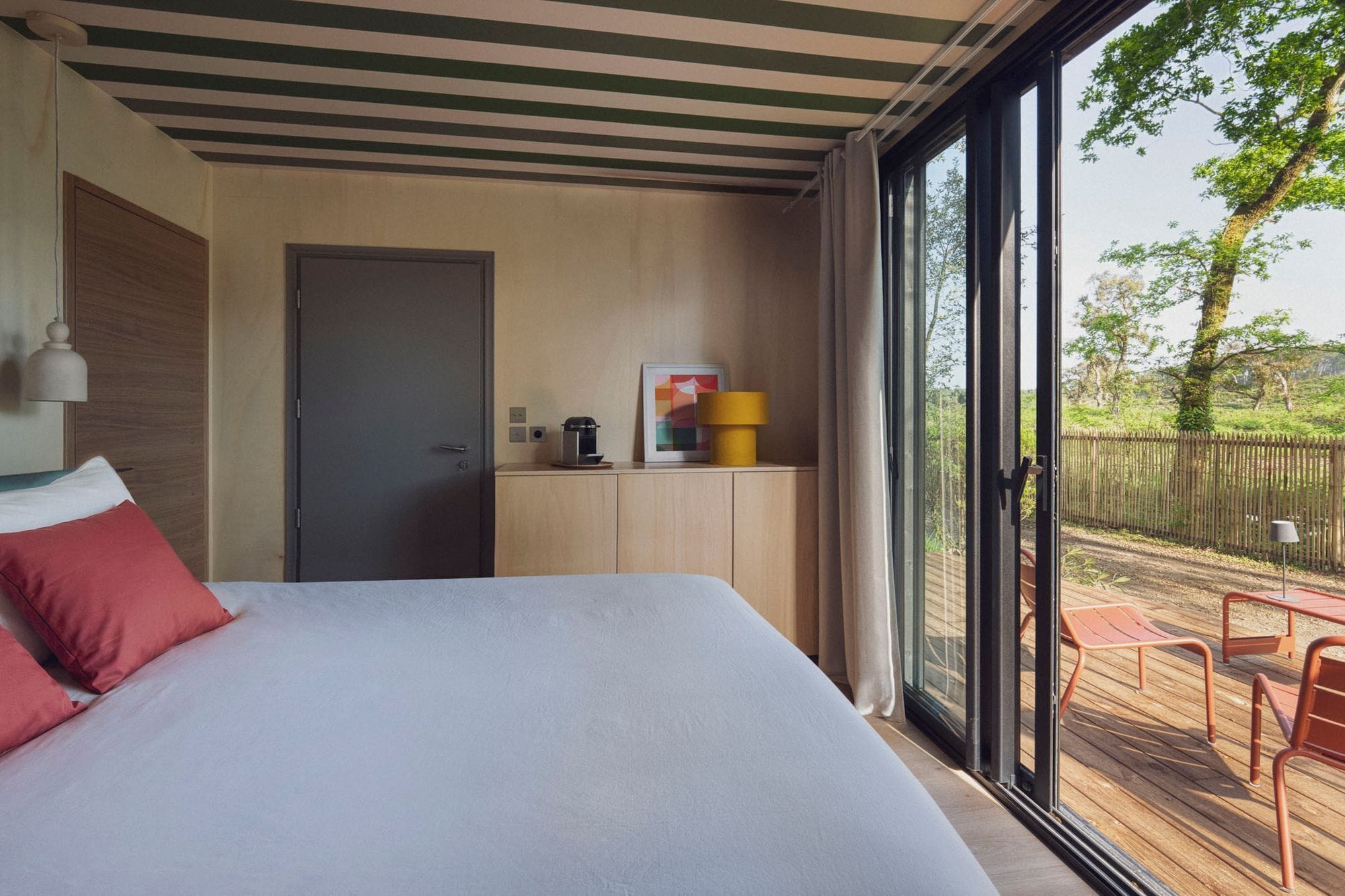 A bedroom with a bed, a door, a small cabinet, and a view of an outdoor deck.