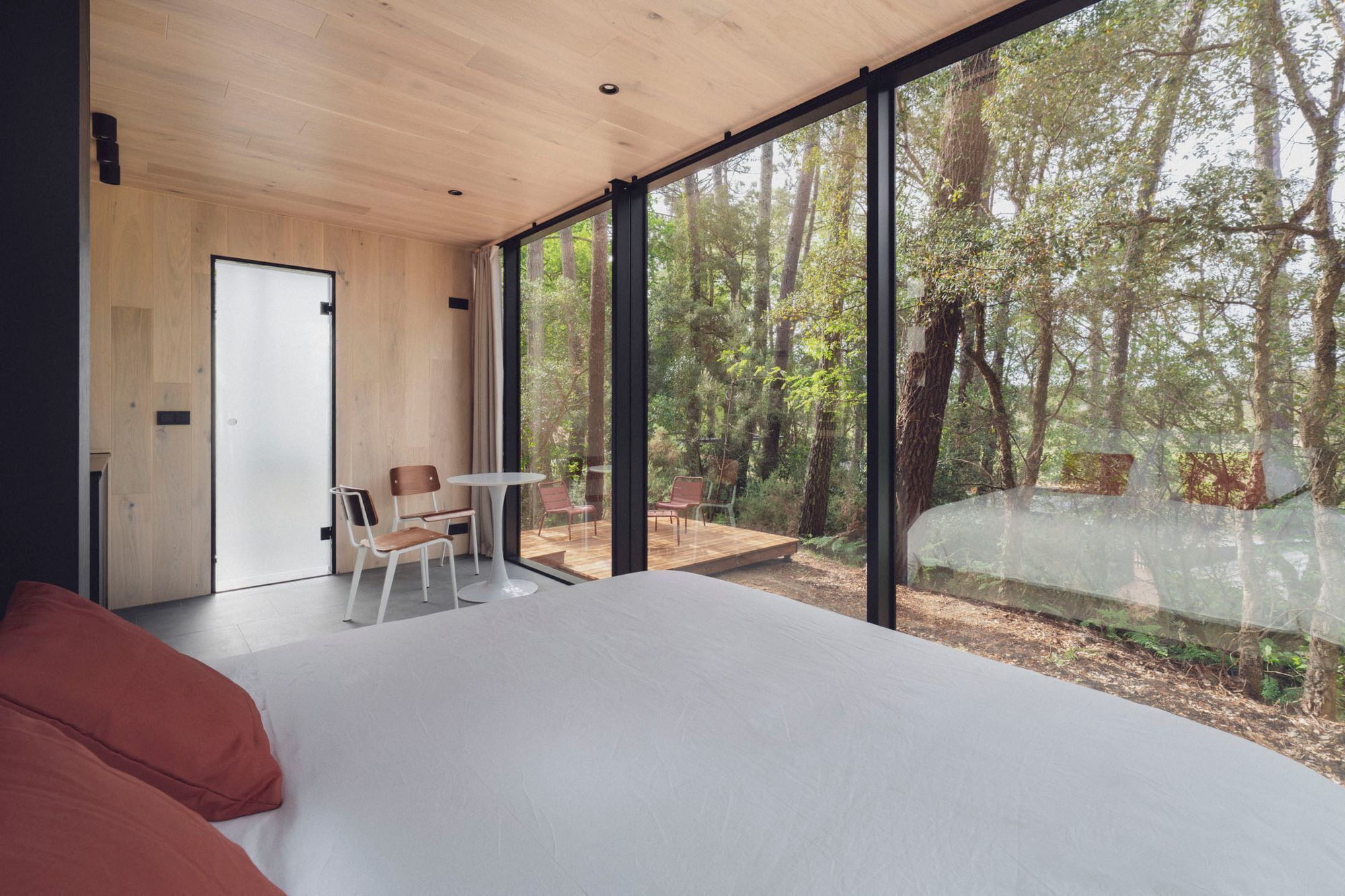 Un dormitorio moderno con una cama grande, una mesita pequeña y sillas, y vistas a un bosque a través de ventanas amplias.