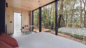 Un dormitorio moderno con una cama grande, una mesita pequeña y sillas, y vistas a un bosque a través de ventanas amplias.