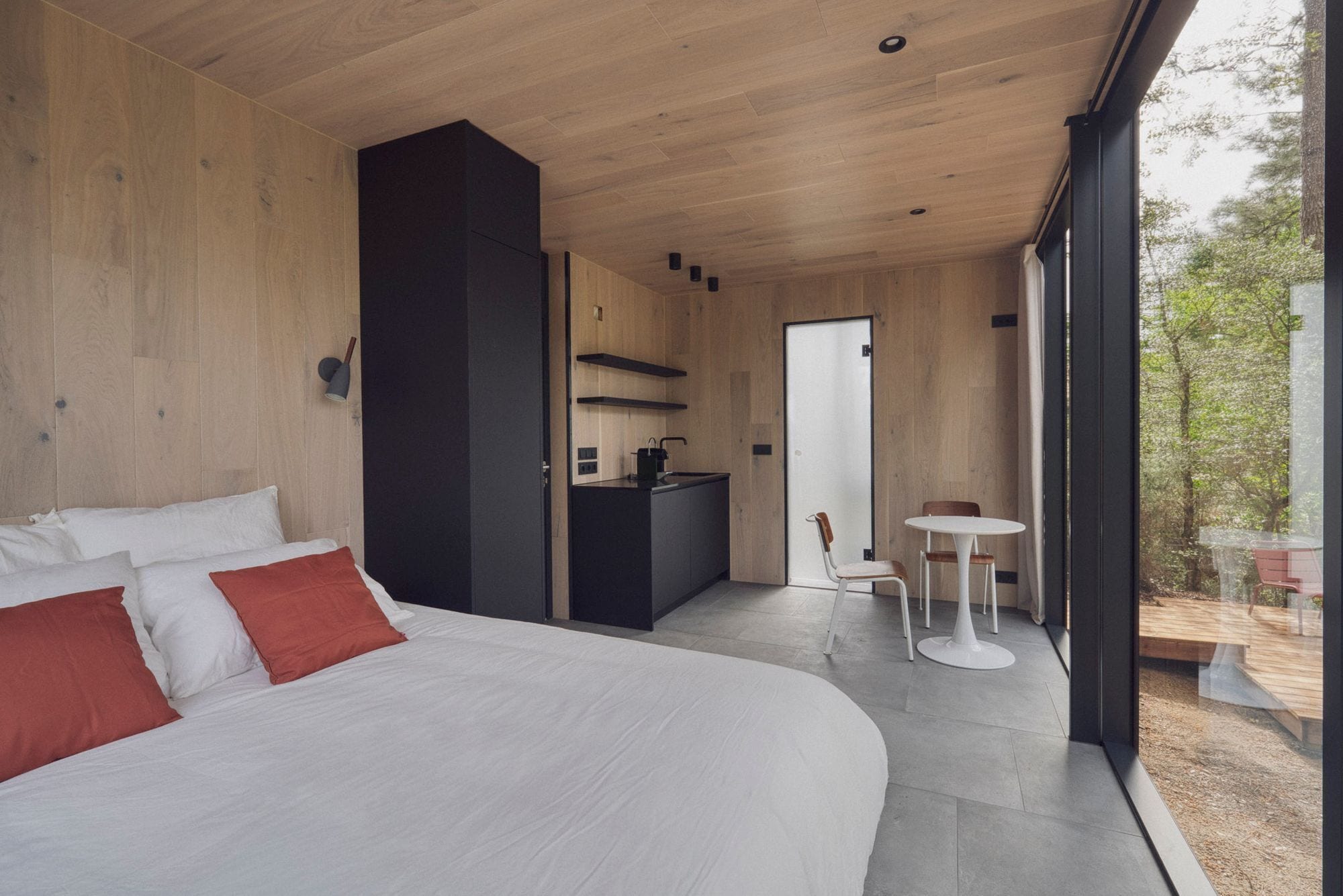 Un dormitorio con una cama grande, un escritorio y una silla, una pequeña cocina y un balcón con vistas a árboles.