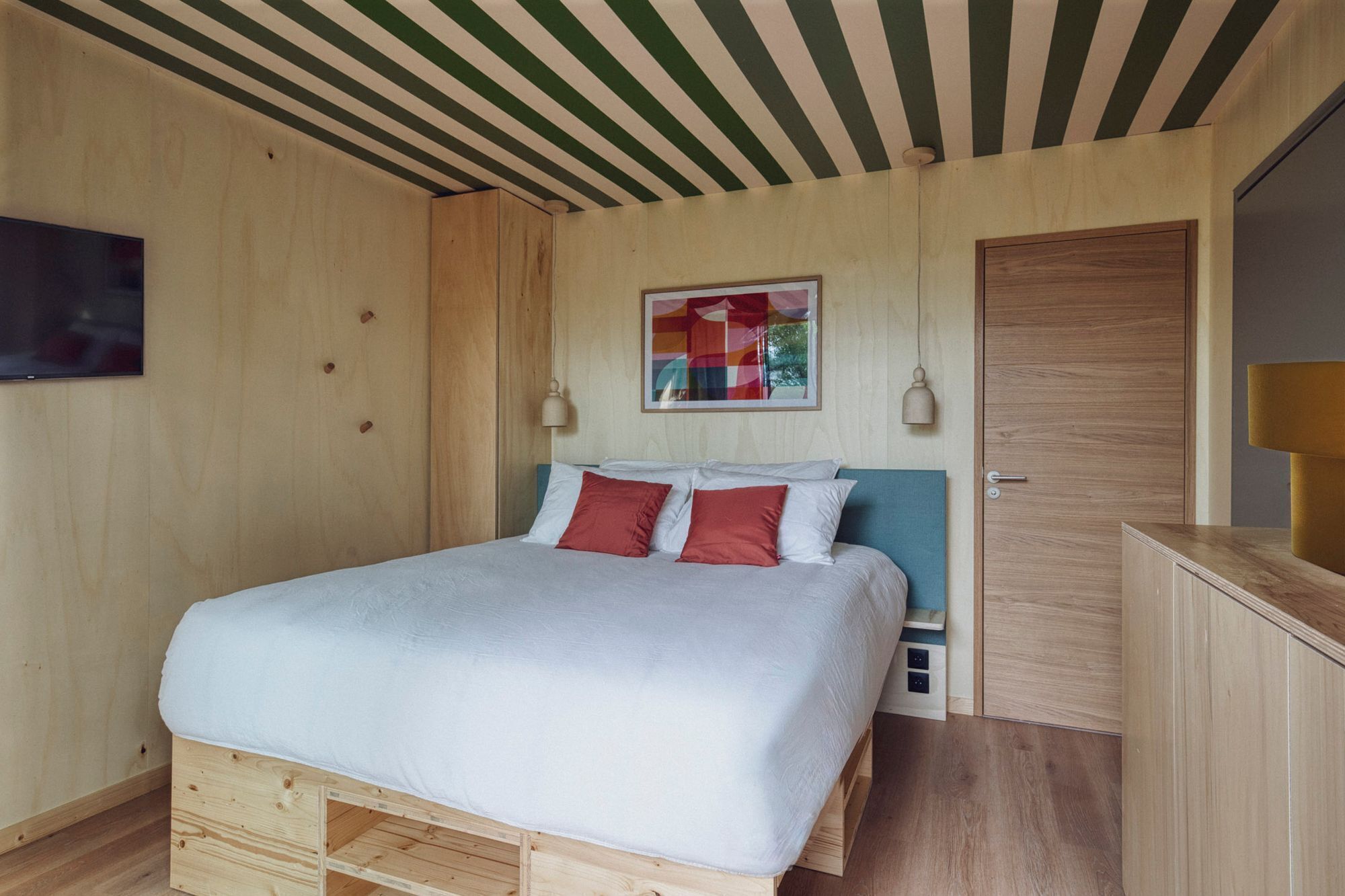 Um quarto com painéis de madeira, uma cama, uma porta e uma TV fixada na parede.