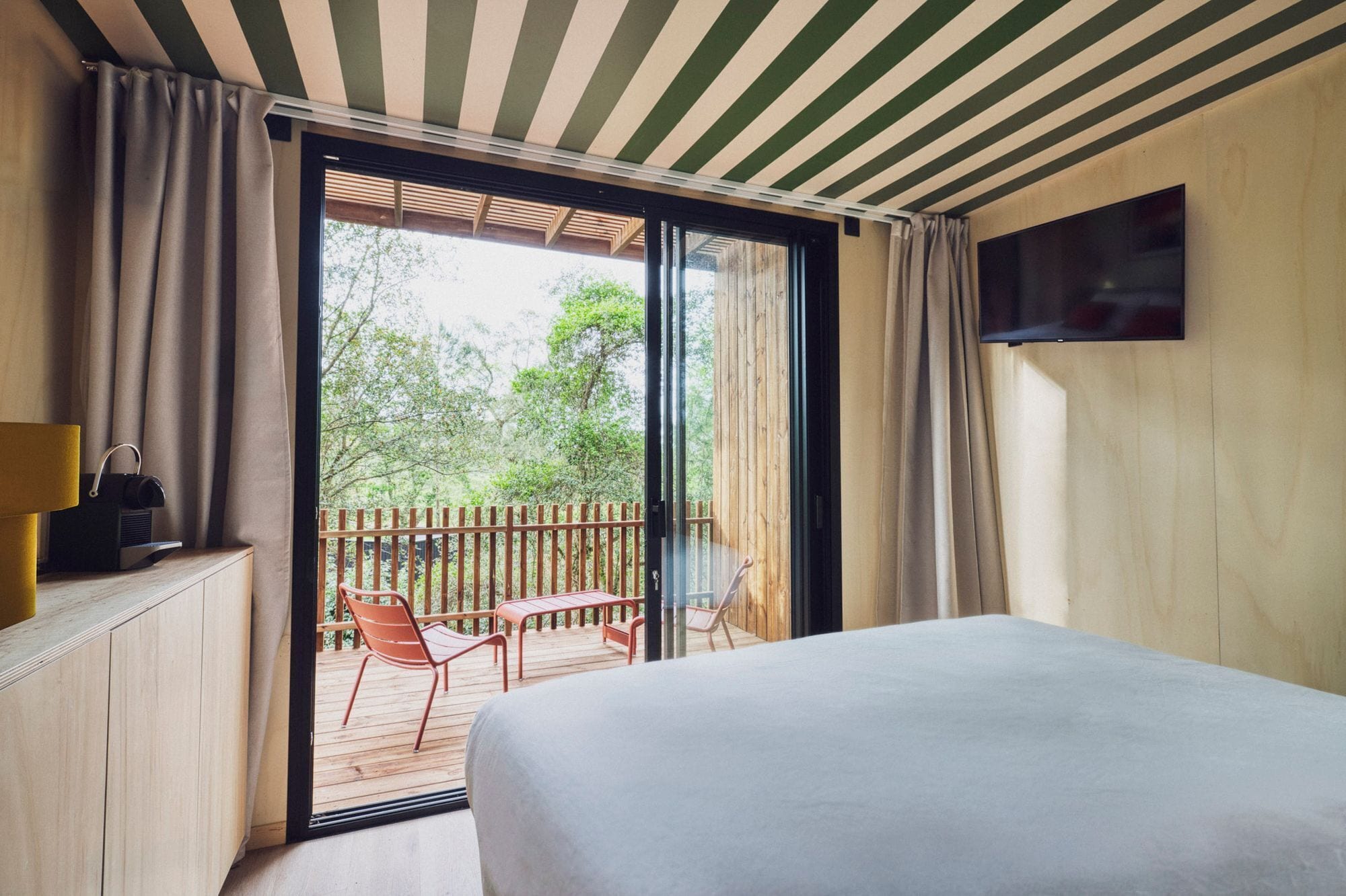 A bedroom with a large bed, a TV mounted on the wall, a wooden desk with a lamp, and a balcony with a table and chairs.