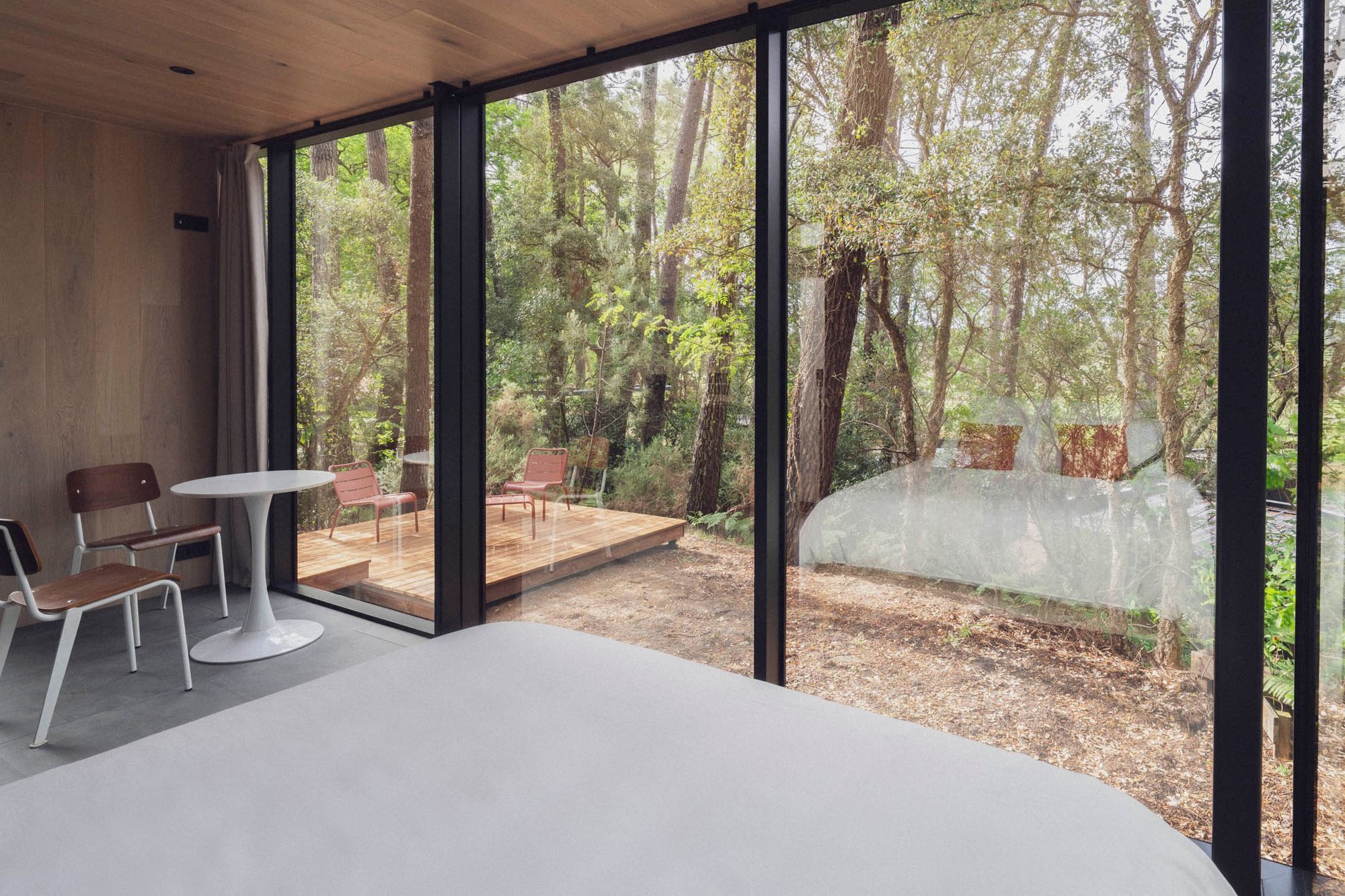 Quarto moderno com amplas janelas de vidro, um deck de madeira e uma área de estar com cadeiras e uma mesa.