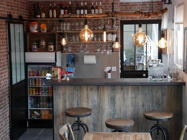 A bar area with wooden stools, shelves with bottles and glasses, and a brick wall.