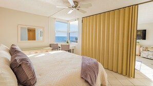 Un dormitorio con una cama grande, un ventilador de techo, un escritorio con silla y vistas al océano a través de una ventana.