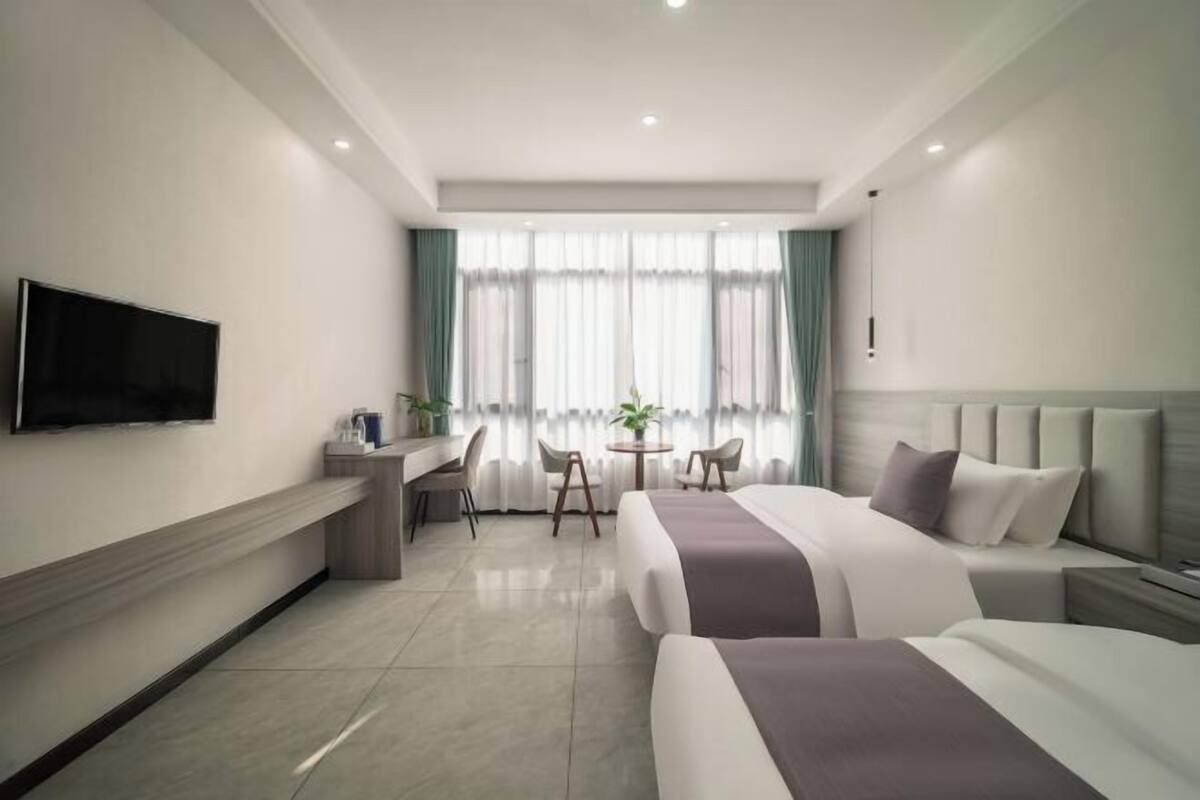 A modern hotel room with a flat-screen TV, a desk with a chair, a round table with chairs, and two beds with headboards.