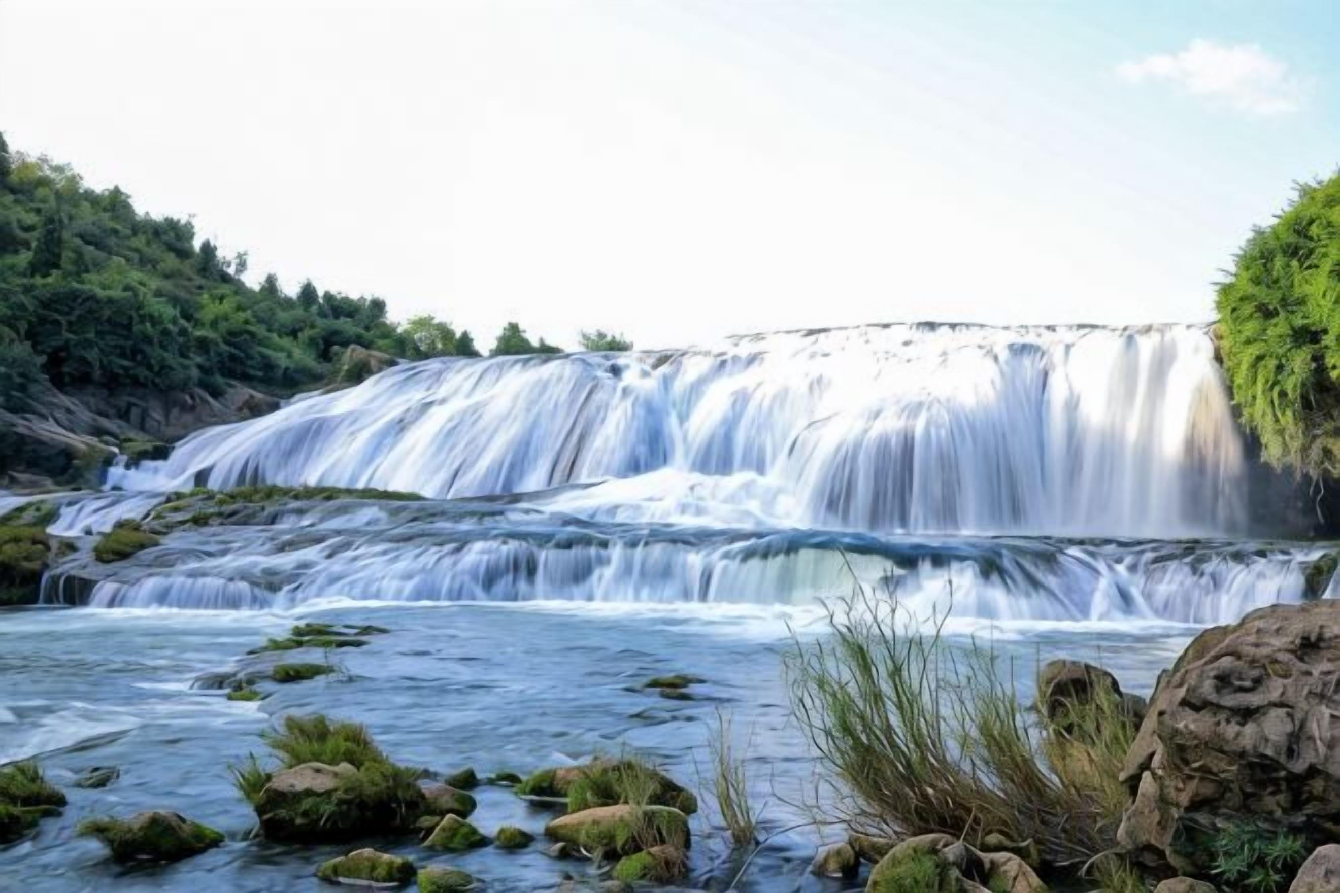 Ein Wasserfall mit felsigem Vordergrund und bewaldetem Hintergrund.