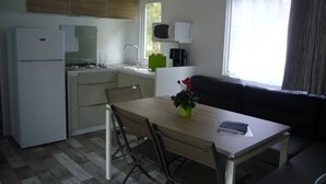 Cocina compacta con refrigerador, estufa y microondas. Hay una mesa de comedor con sillas y una pequeña planta en maceta. Se ve un sofá en la zona de estar contigua.