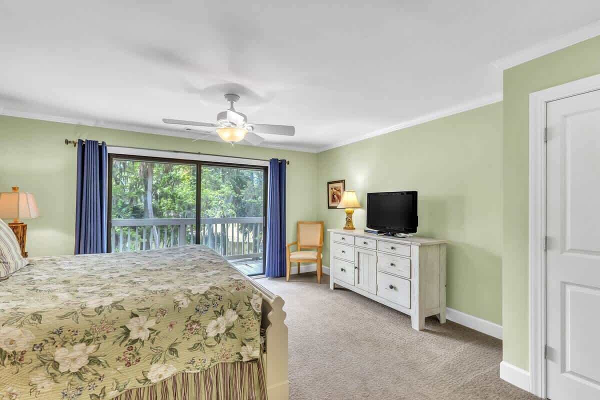 Ein Schlafzimmer mit einem Bett, einem Fernseher, einem Deckenventilator, einem Fenster mit Vorhängen und einem Balkon.