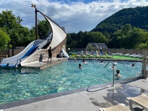A swimming pool with a slide, surrounded by a wooden deck and a mountain backdrop.