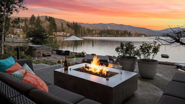 Eine Terrasse mit Feuerstelle, Gartenmöbeln und Blick auf einen See und Berge bei Sonnenuntergang.