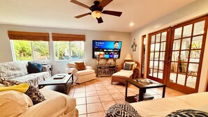 Un salón con sofá, sillón, reposapiés, televisor y un ventanal con persianas.