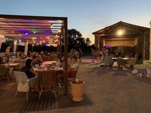 Un espace extérieur aménagé pour les repas, avec des tables et des chaises, une pergola illuminée de guirlandes lumineuses et, au fond, un bâtiment au style rustique.