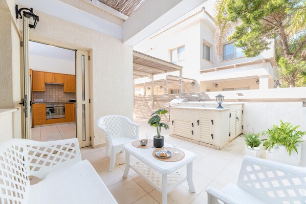 Une terrasse extérieure lumineuse, avec des meubles blancs, une petite table agrémentée d’une plante en pot et une vue sur la cuisine.
