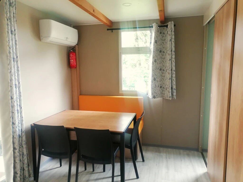 Una habitación pequeña con una mesa y sillas de madera, una ventana con cortina y un equipo de aire acondicionado.