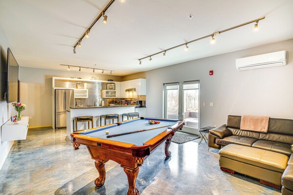 Un salon moderne avec une table de billard, un canapé et un coin cuisine à l’arrière-plan.