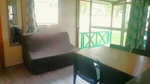 A room with a sofa, a table, and a chair. There is a window with curtains and a view of a green deck outside. - ZEcamping 4 étoiles piscine dog friendly animations chalet TV BBQ clim (Saint-Gaultier)