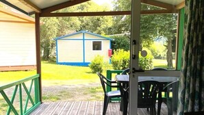 A covered outdoor area with a wooden deck, a table with chairs, and a small building in the background. - ZEcamping 4 étoiles piscine dog friendly animations chalet TV BBQ clim (Saint-Gaultier)