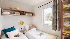 Une chambre avec deux lits, une fenêtre avec des rideaux, une étagère avec des serviettes pliées et une petite étagère ronde avec des livres.