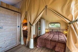 Una habitación con tienda de campaña acogedora, con suelo de madera, una cama con colcha marrón y una ventana que ofrece vistas al follaje exterior.