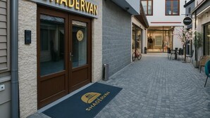 A hotel entrance with a number 24 door, a doormat with the hotel name, and a cobblestone pathway leading to a seating area with chairs and a bicycle. - Hotel Shadervan (Prizren)