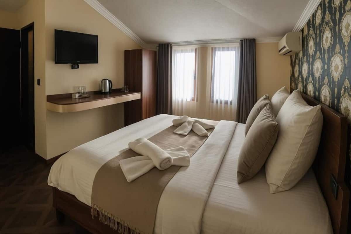 A hotel room with a large bed, a flat-screen TV mounted on the wall, a small table with a kettle, and a patterned wallpaper on the wall.