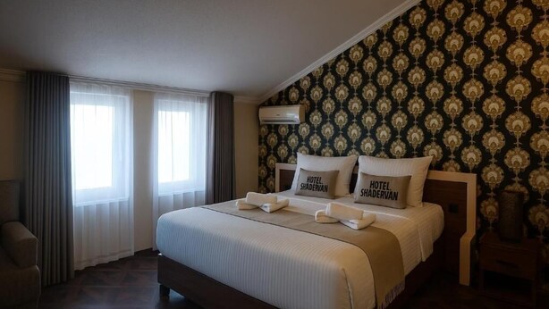 A hotel room with a large bed, two pillows with the hotel's name, a nightstand, and a window with curtains. - Hotel Shadervan (Prizren)