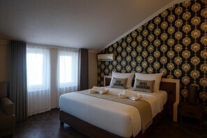 Habitación de hotel con una cama grande, dos almohadas con el nombre del hotel, una mesita de noche y una ventana con cortinas.