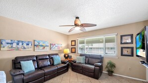 A living room with a brown leather sectional sofa, a ceiling fan, a flat-screen TV, and a window with blinds.