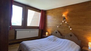 Ein Schlafzimmer mit einem Holzkopfteil, einem großen Bett mit weißer Bettdecke, einem Fenster mit roten Vorhängen und einer Wandlampe.