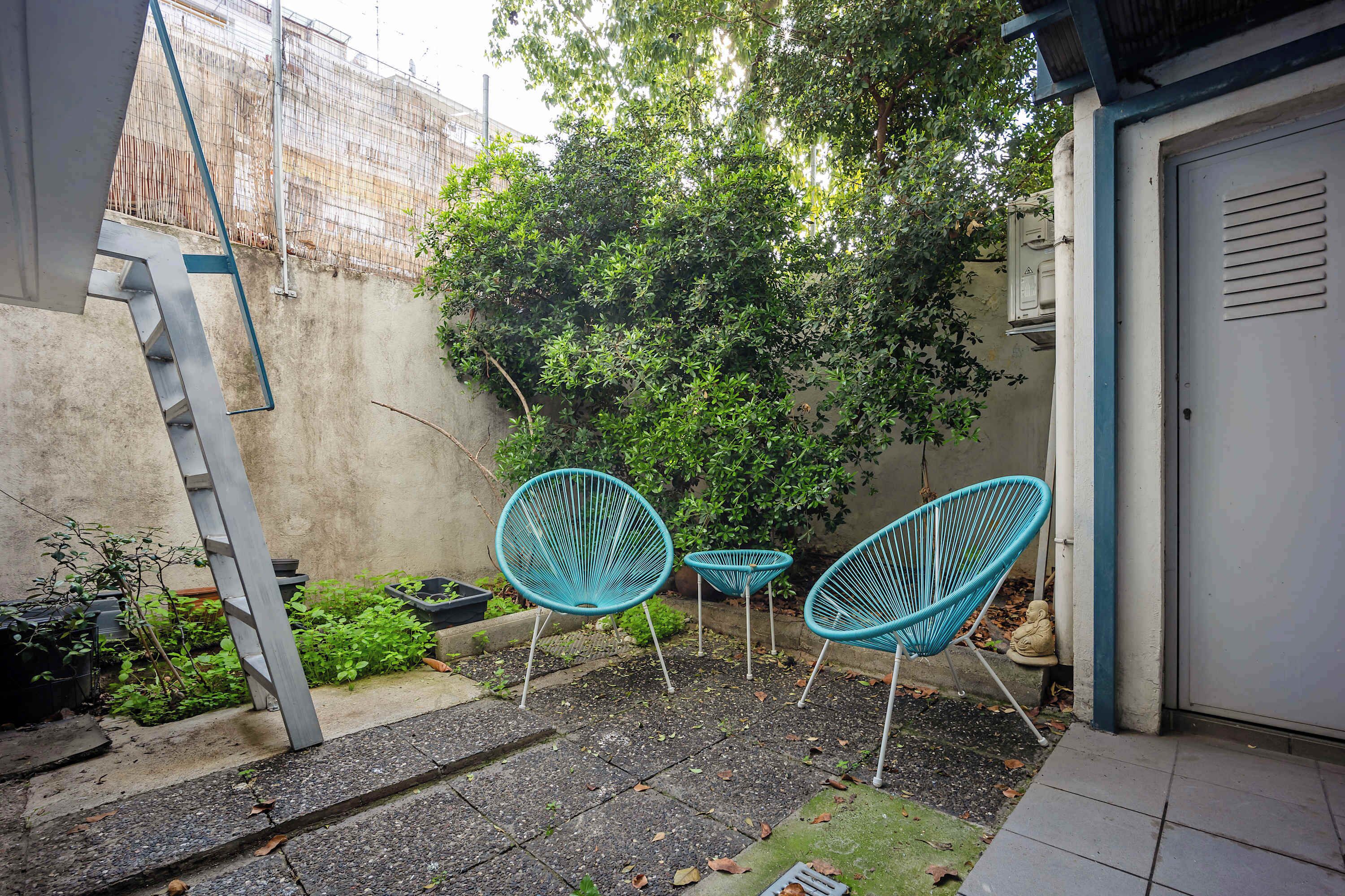 A small outdoor patio area with three turquoise chairs, a concrete floor, and a metal ladder leaning against a wall.