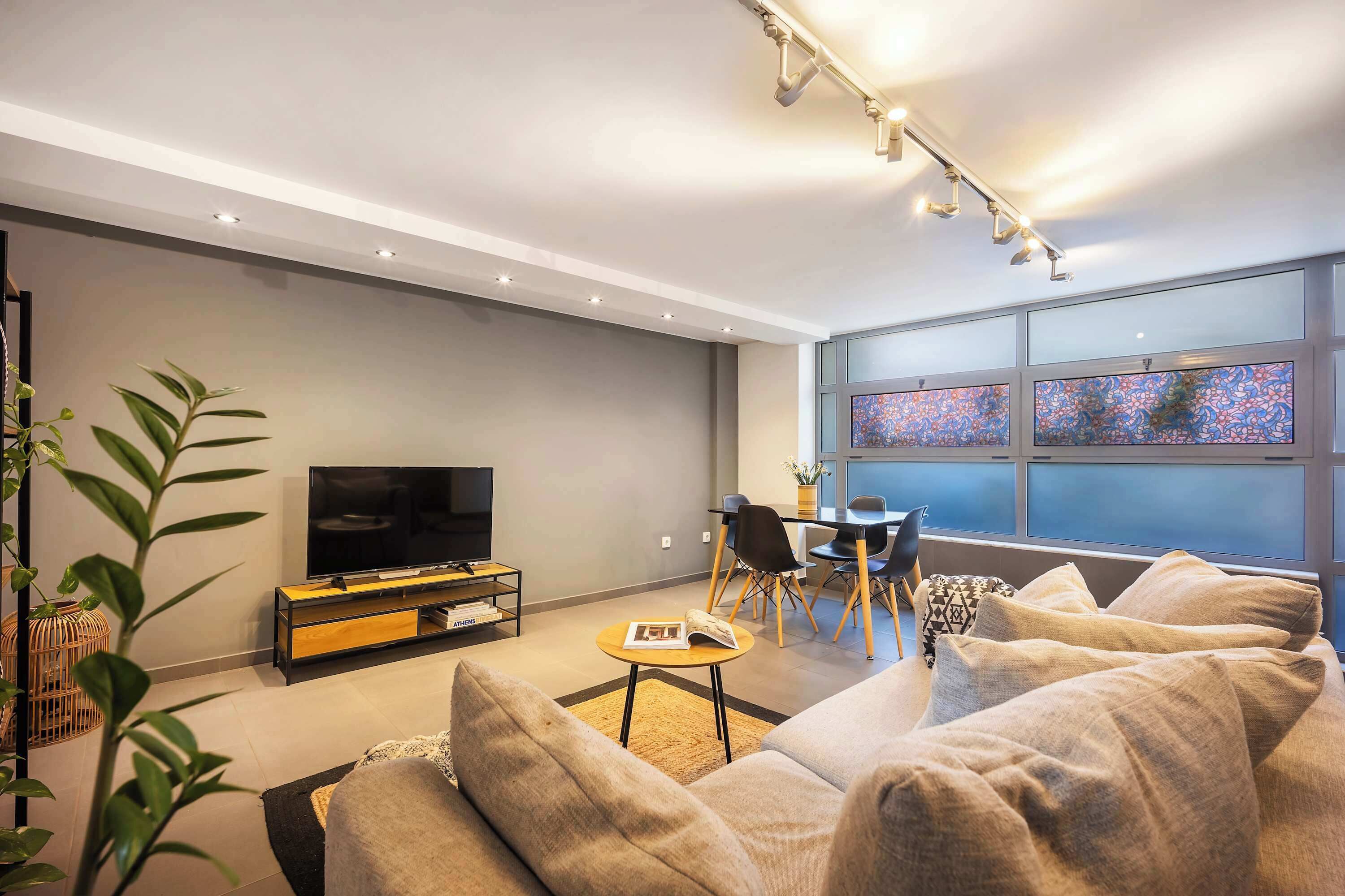 A modern living room with a flat-screen TV, comfortable seating, a dining area, and large windows with decorative glass panels.