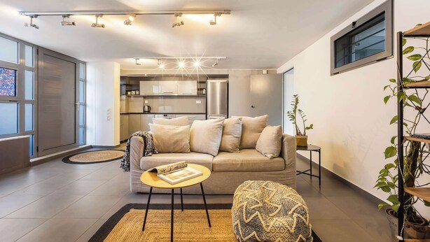 Ein modernes Wohnzimmer mit einer Couch, einem Couchtisch und einem Teppich. Im Hintergrund ist eine Küche mit Schränken und einem Kühlschrank zu sehen.