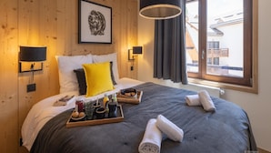A modern hotel room with wooden paneling, a large bed with a gray and black duvet, a framed artwork on the wall, and a window with curtains. - Chalet in Serre Chevalier With Family Fun (Saint-Chaffrey)