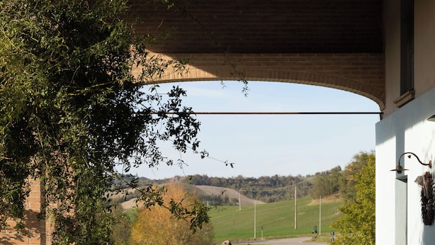 An outdoor patio with a wooden bench, a table, and chairs. A view of a rural landscape with a road and trees is visible through an arched structure.