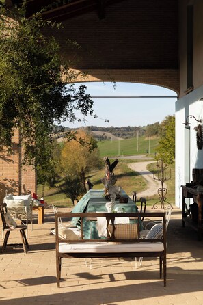 An outdoor patio with a wooden bench, a table, and chairs. A view of a rural landscape with a road and trees is visible through an arched structure. - AGRITURISMO CASA DELLE VIOLE (Emilia-Romagna)