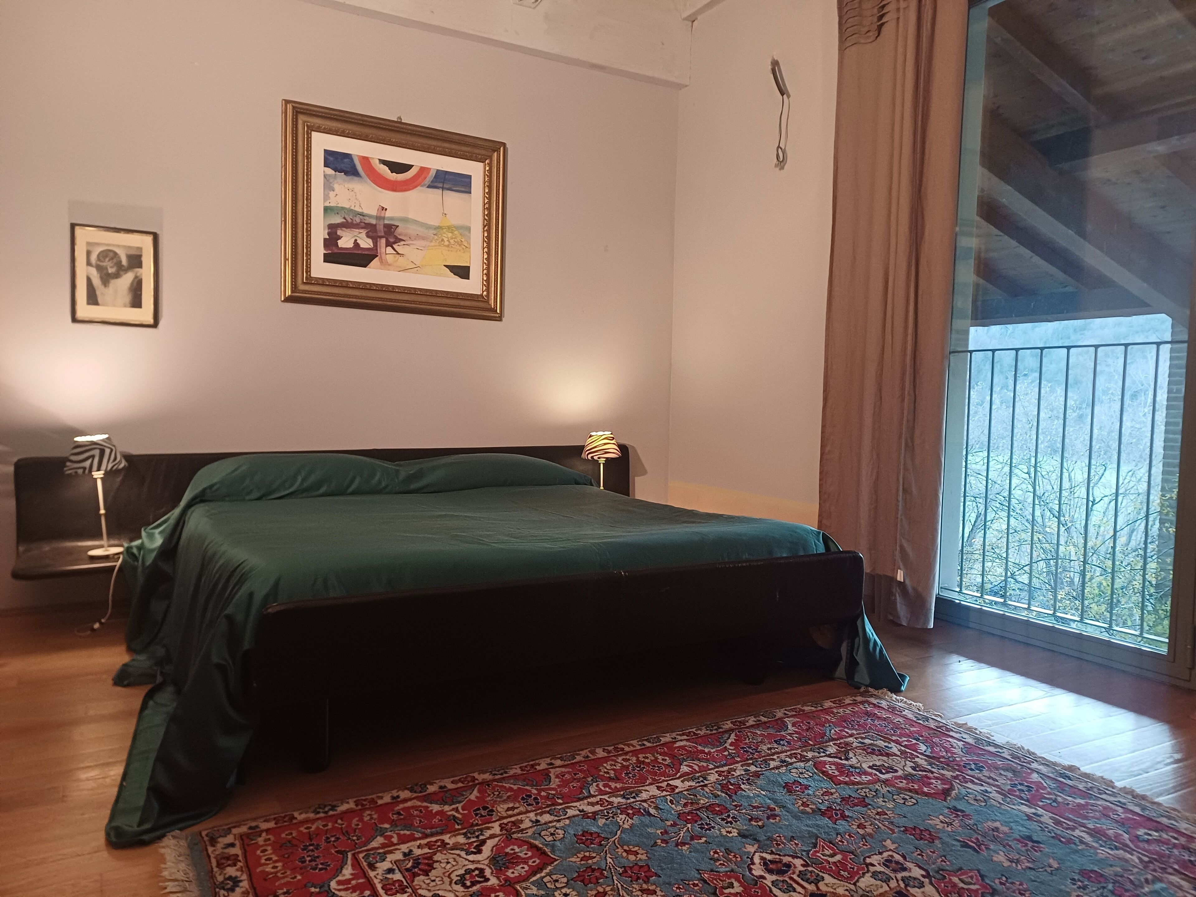 Una camera da letto con un letto, un comodino, una lampada, un quadro incorniciato e una finestra vista alberi.