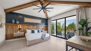 A bedroom with a large bed, wooden ceiling beams, a ceiling fan, a nightstand, a bedside table, and a view of the outdoors through a sliding glass door.