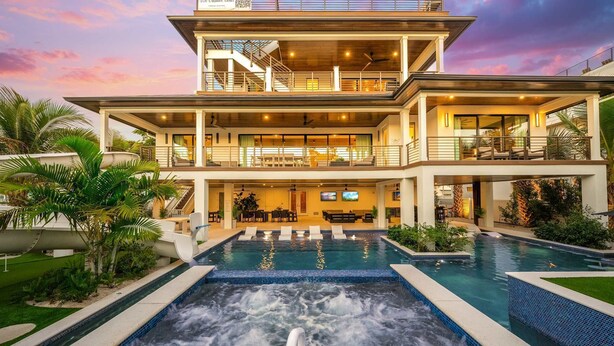 A multi-story house with a large pool, outdoor seating, and a staircase leading to a balcony.