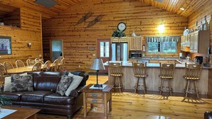 L’intérieur chaleureux d’un chalet en bois comprend un coin repas, un salon avec un canapé en cuir, une cuisine avec des meubles en bois et une grande horloge au mur.