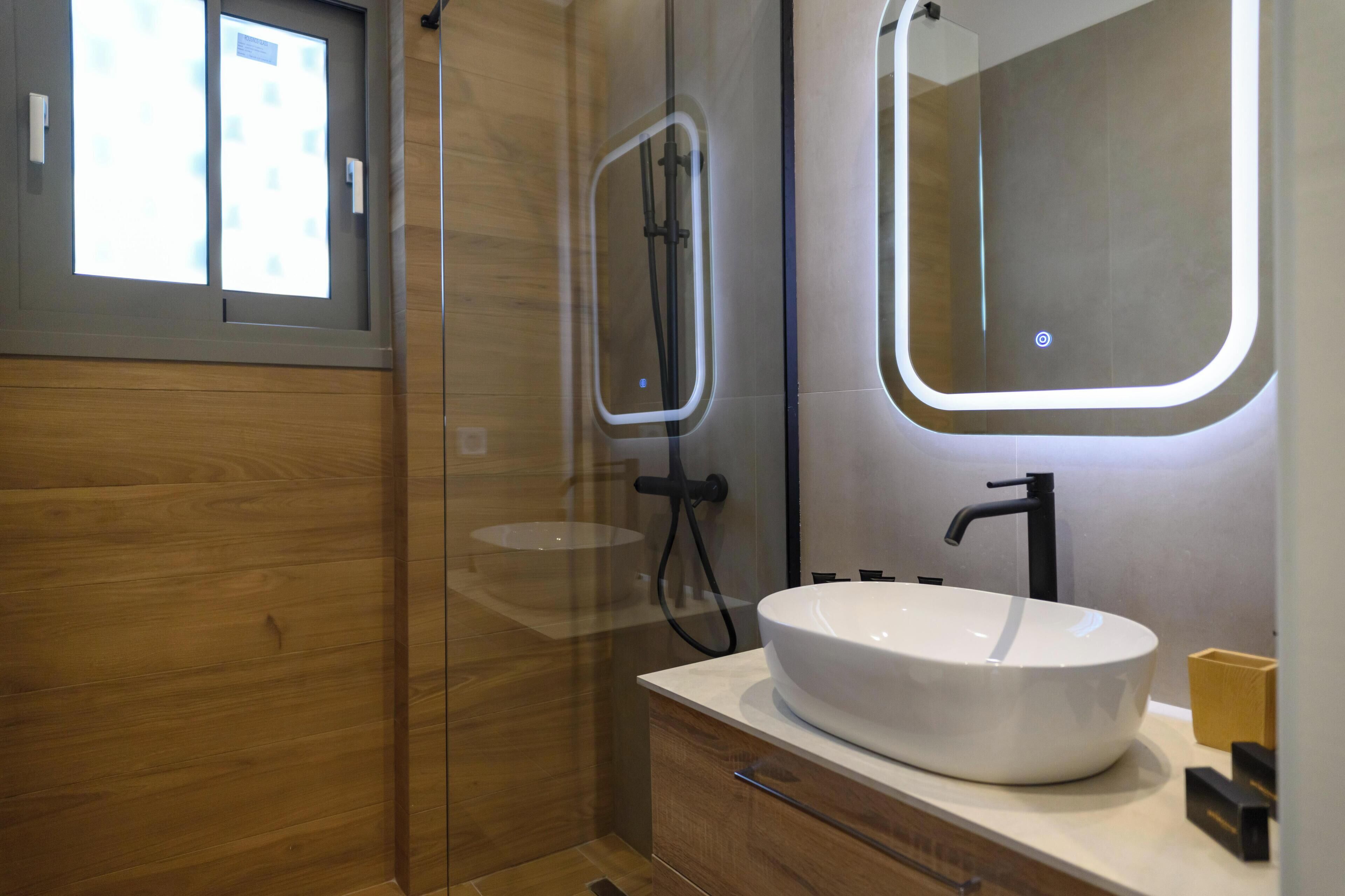 Un baño moderno con cabina de ducha de vidrio, un lavabo blanco con grifo negro y un espejo con marco iluminado.