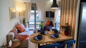 A modern living room with a sofa, dining table, and chairs. There is a TV mounted on the wall, a refrigerator, and a window with curtains.